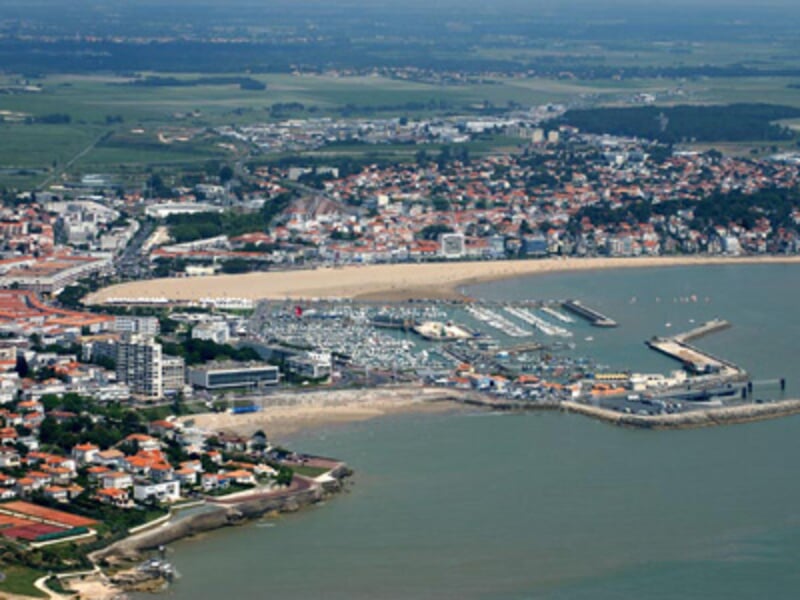 Vue aérienne d'une ville côtière avec une plage, un port de plaisance et des bâtiments résidentiels environnants.